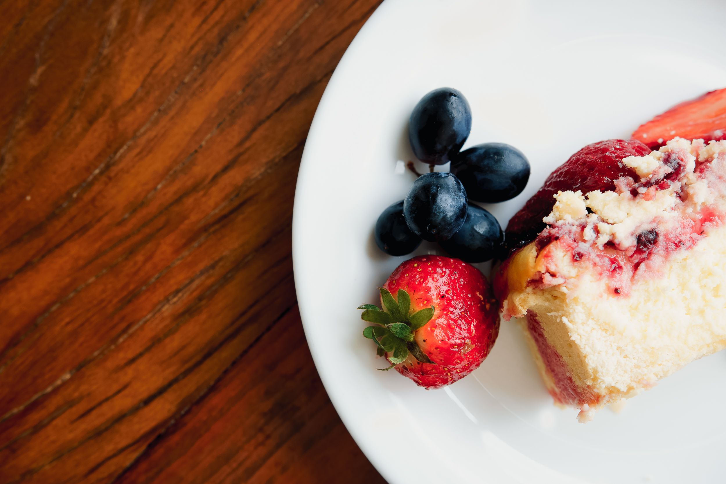 Indulgent slice of cheesecake with strawberries and grapes on a wooden table.