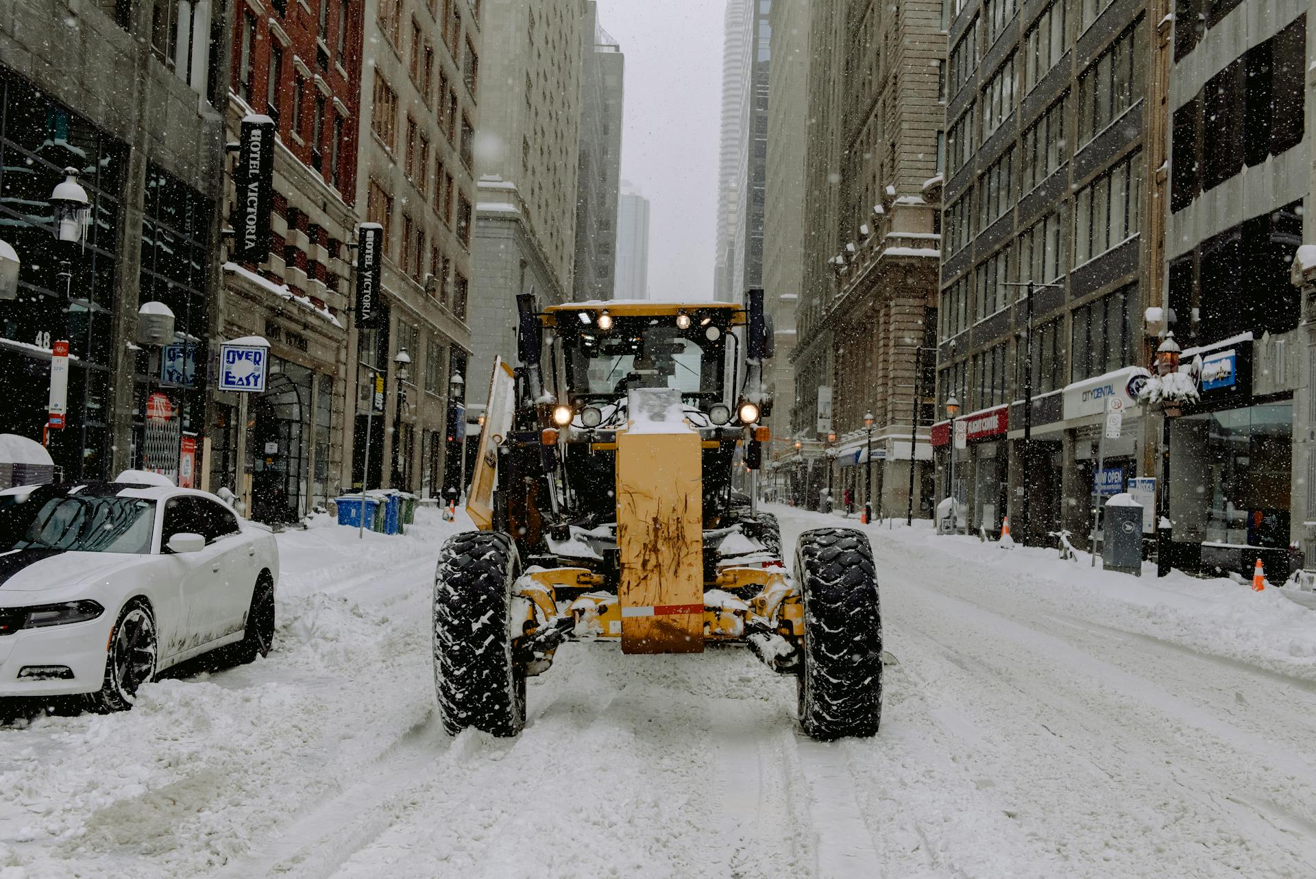 A motor grader clearing snow on a city street during heavy snowfall, winter urban scene.