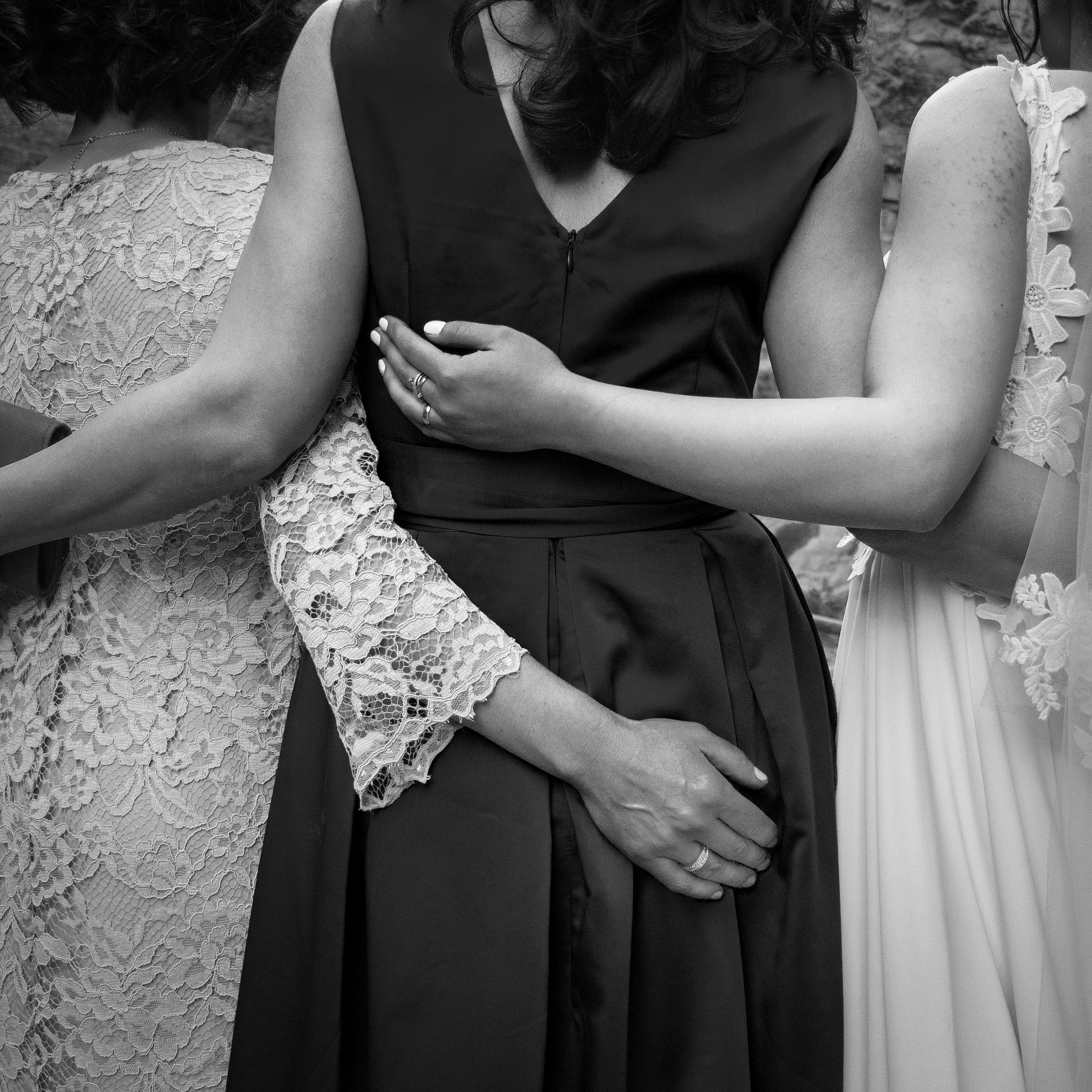 Three women in formal dresses embrace, showcasing togetherness in a black and white photo.