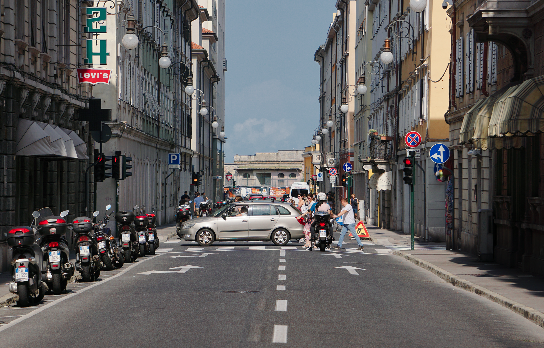 A bustling street scene in downtown Trieste, Italy, featuring traffic, pedestrians, and historic architecture.
