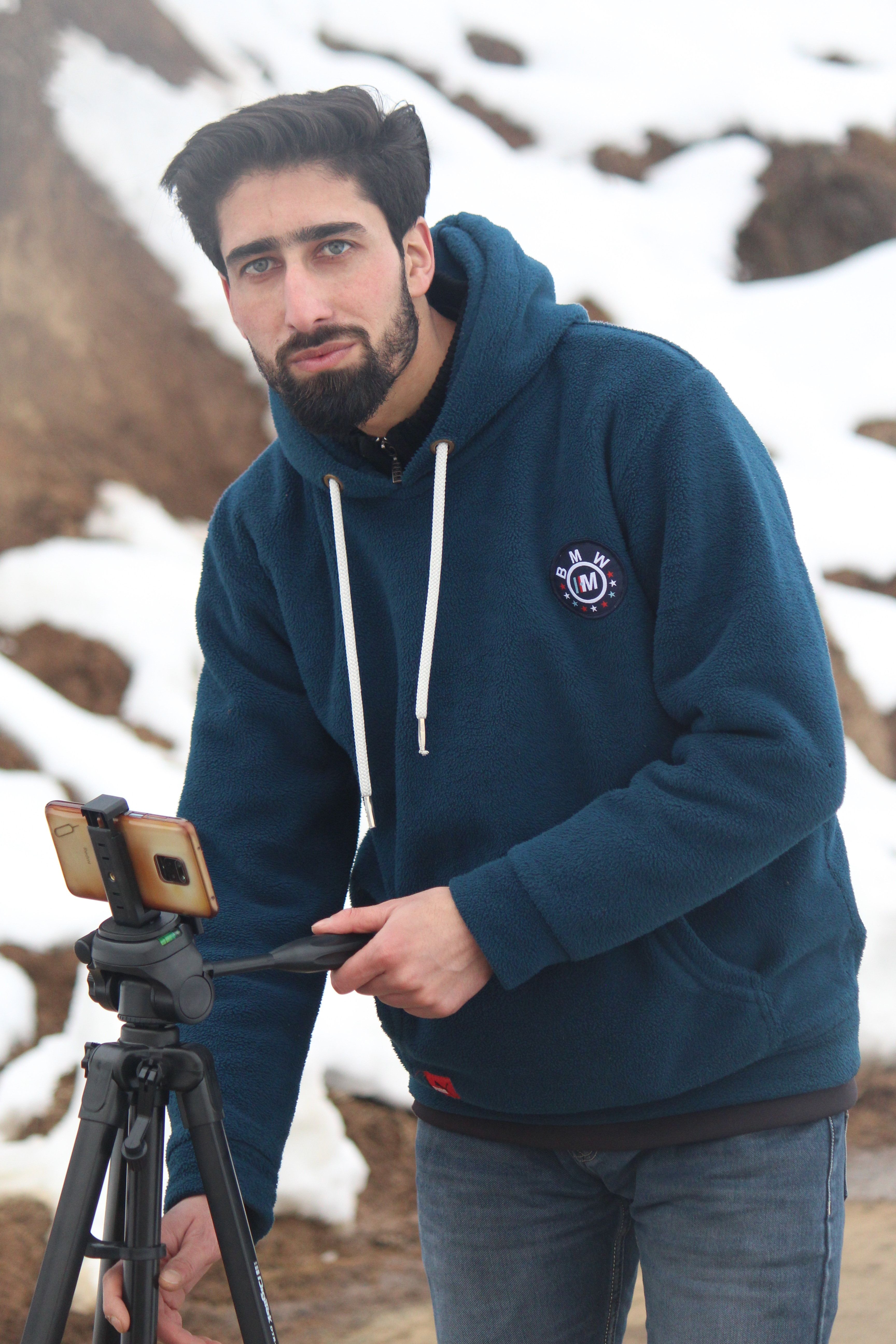 A young man in a blue hoodie operates a tripod-mounted smartphone outdoors in snowy winter scenery.