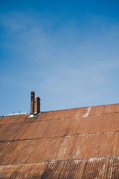 Free Rustic corrugated metal roof with a chimney against a clear blue sky, conveying simplicity and rural charm. Stock Photo