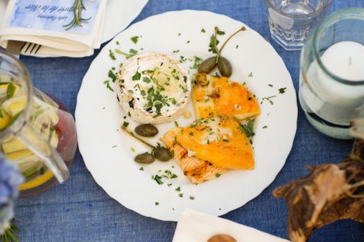 Flat lay of a gourmet meal with cheese, grilled vegetables, and capers on a blue table setting.