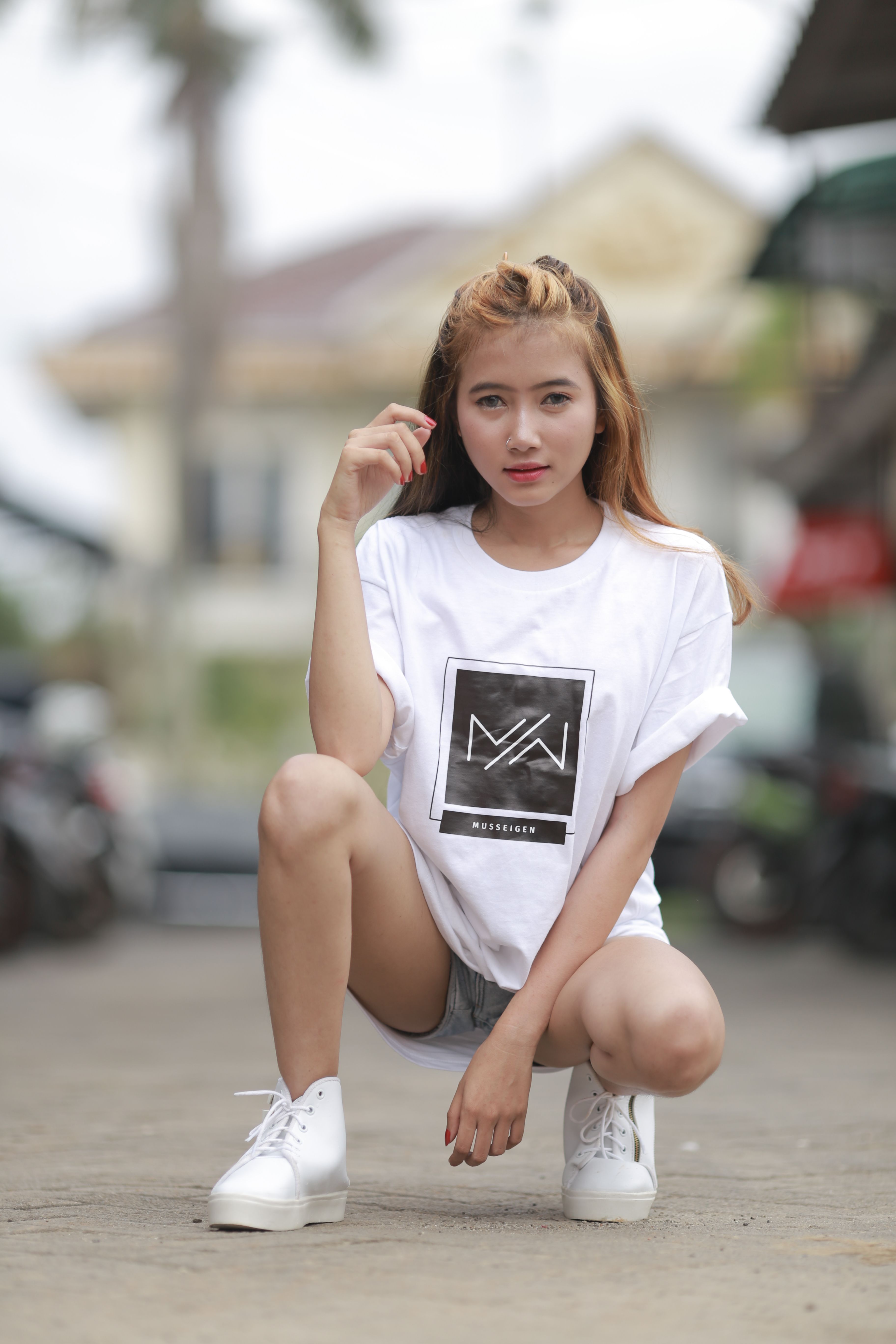 Young woman with long hair squatting outdoors in a white t-shirt and sneakers, showcasing urban style.