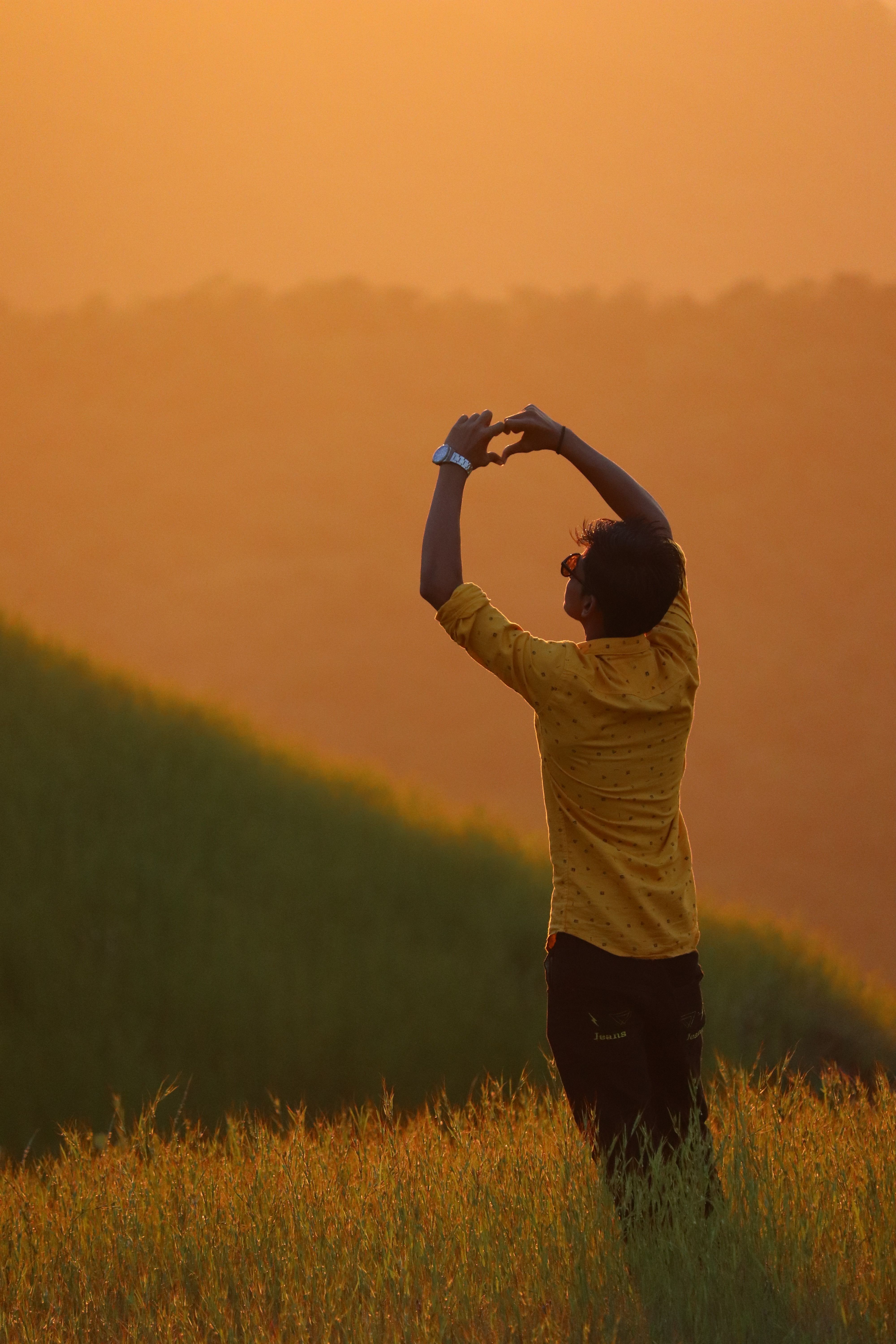 Back view of a man in yellow sleeves forming heart sign during a sunset in a grassy field.