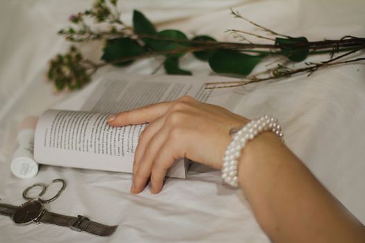 Close-up of a hand with pearl bracelet holding a book, surrounded by flowers and a watch, creating an elegant scene.