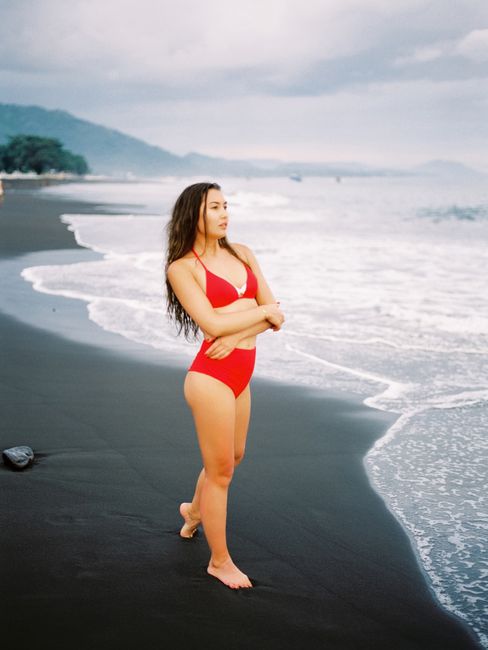 A woman in a red bikini stands on Bali's scenic black sand beach, watching the waves.