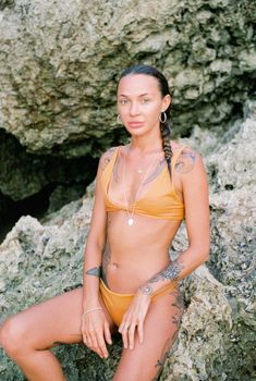 Young tattooed woman in an orange bikini posing against rocky landscape in Bali.