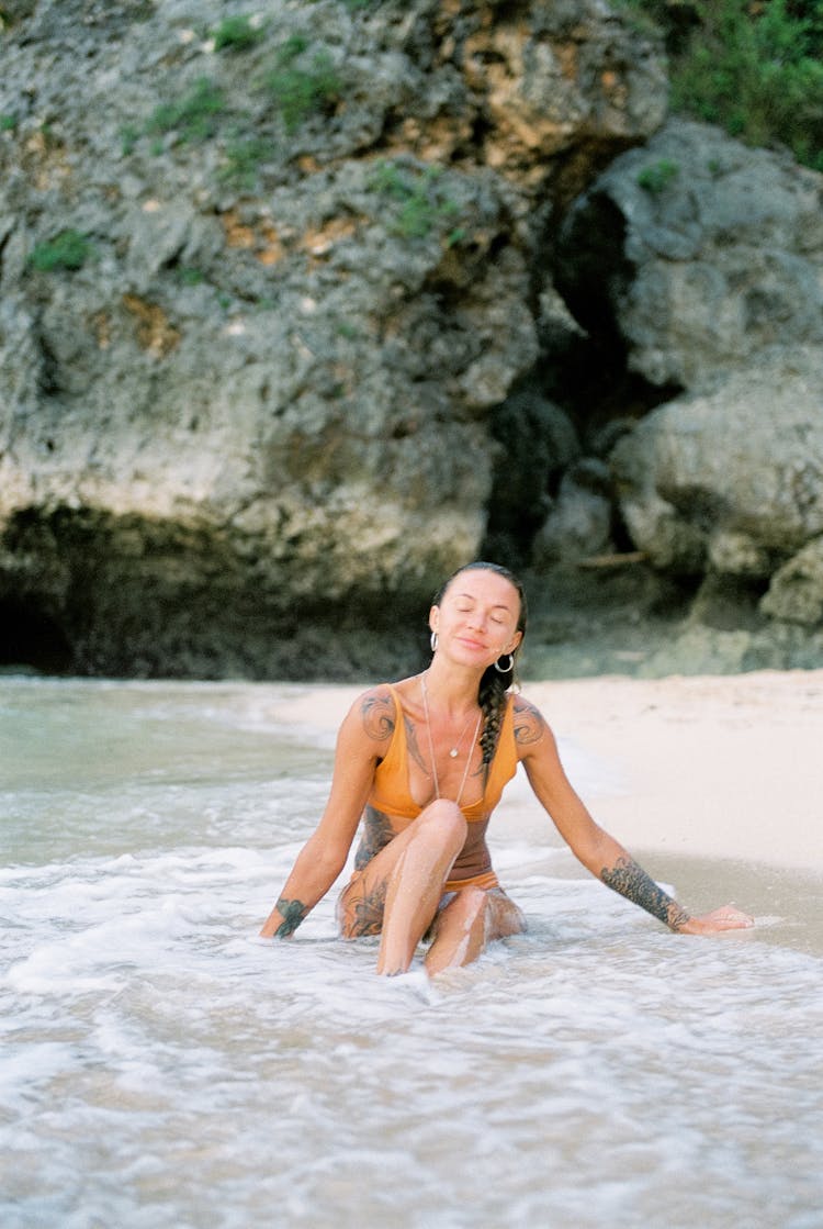 Tattooed Woman In Bikini Sitting With Closed Eyes At Seashore And Enjoying Water