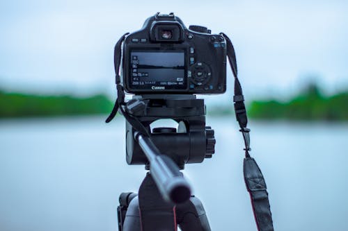 Selective Focus Photo of Black Canon Camera on Tripod Stand in Front of Body of Water Photo