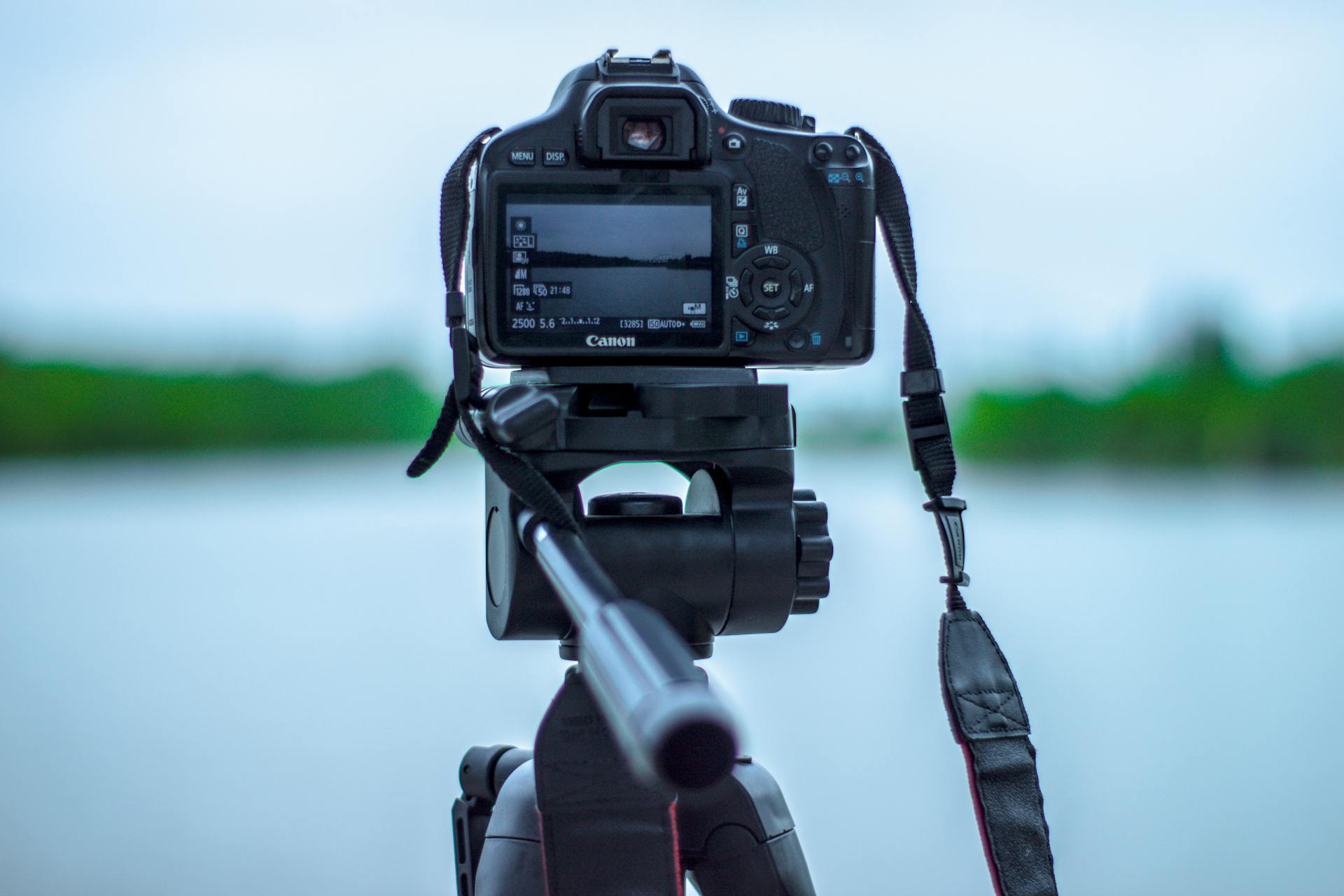 Selective Focus Photo of Black Canon Camera on Tripod Stand in Front of Body of Water Photo