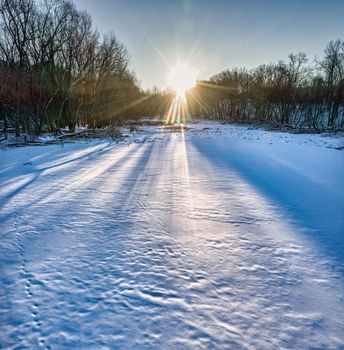A picturesque winter sunrise casting long rays over a snow-covered field and bare trees.