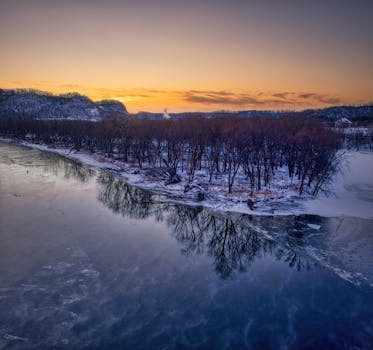 A serene winter sunrise over the Mississippi River with snow-covered trees reflecting in the water.