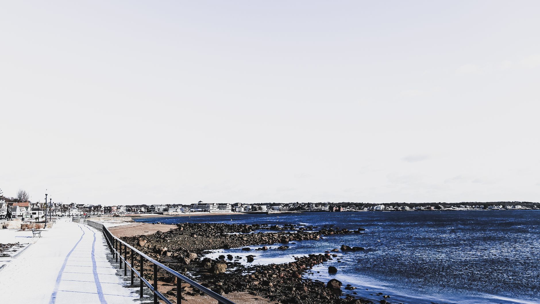 Enjoy a serene winter view of the Manchester-by-the-Sea coastline with snow-covered paths and rocky shore.
