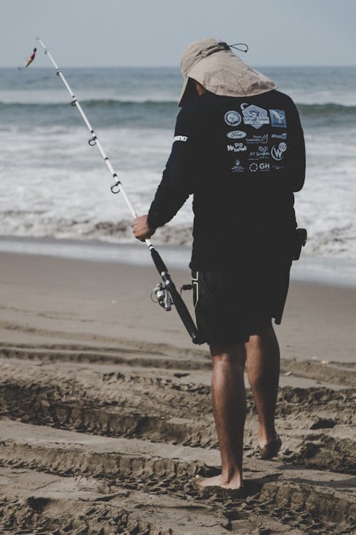 Hombre En La Parte Superior Negra De Manga Larga Con Caña De Pescar Blanca Cerca De La Playa