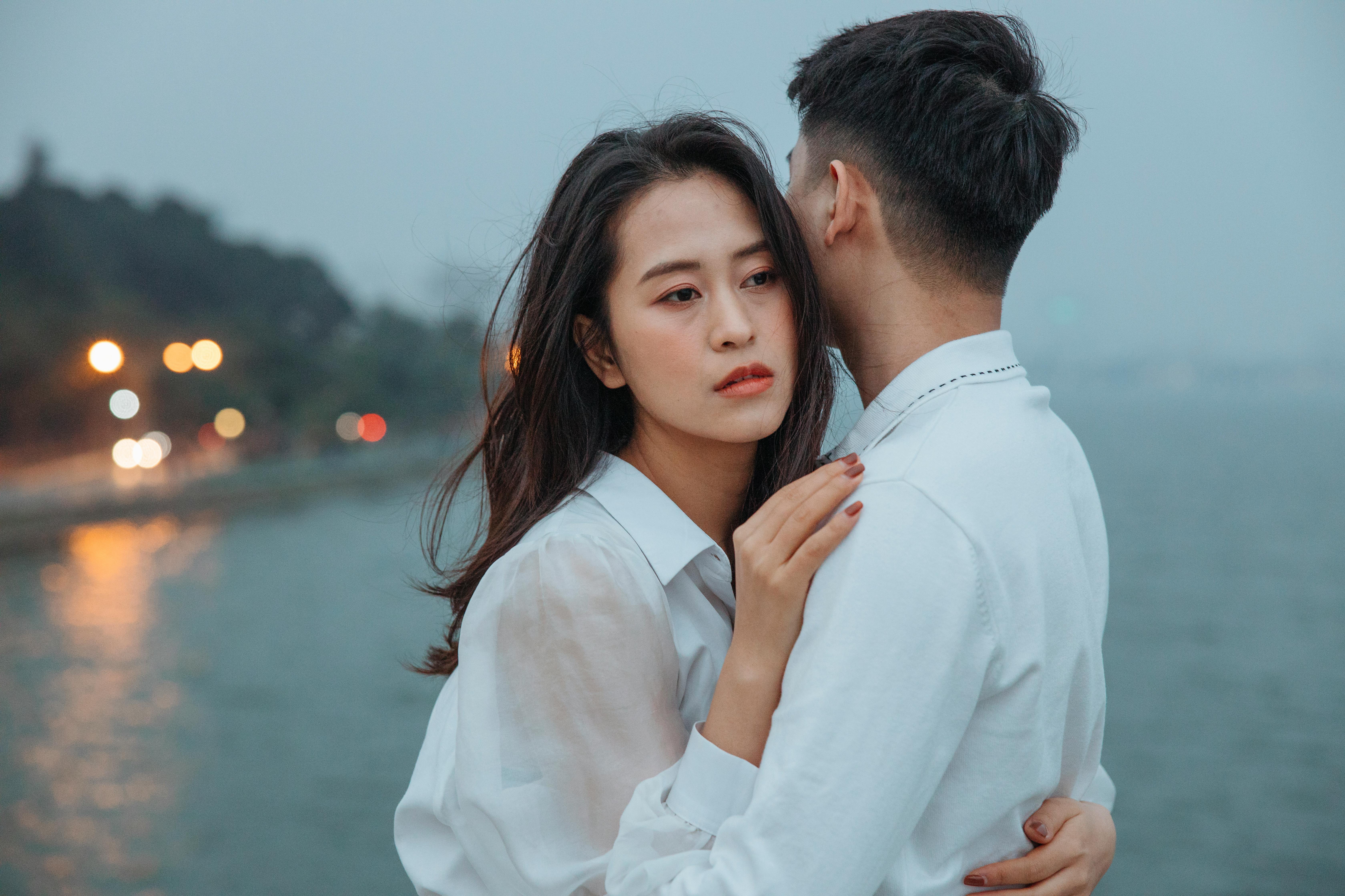 A young couple in white tops embraces near a waterfront, capturing a moment of romance and affection.