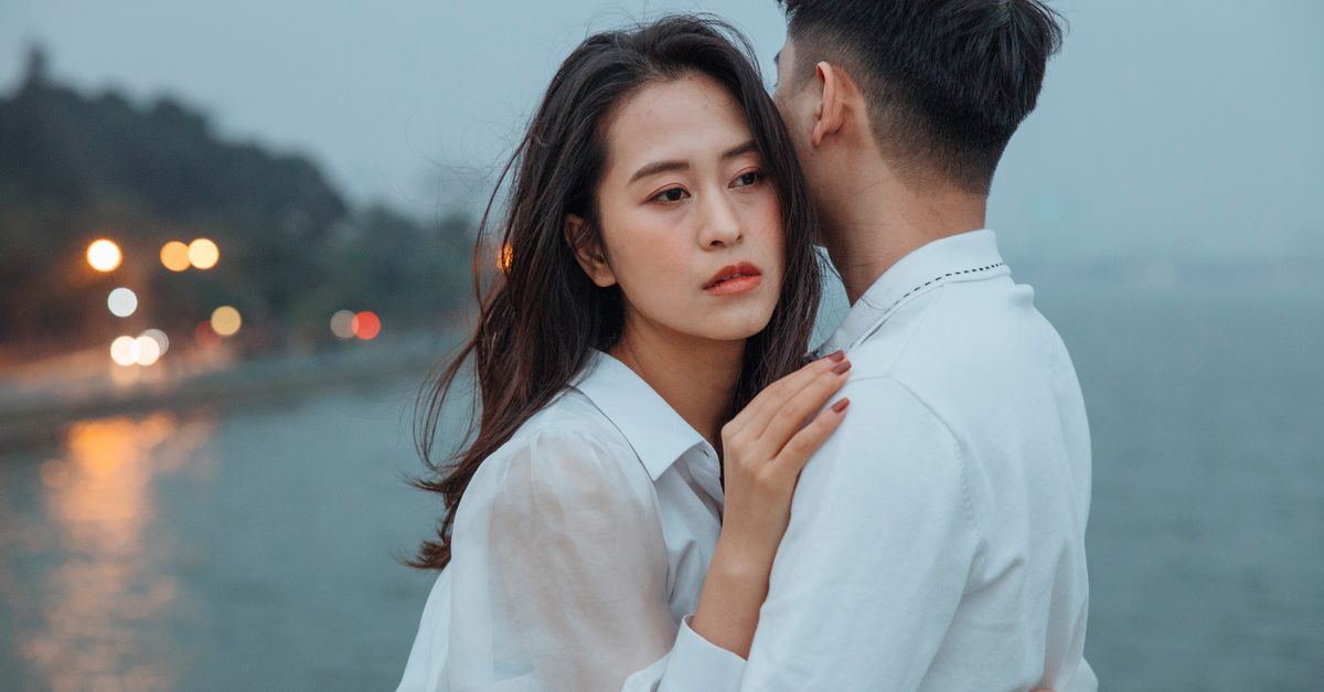 A young couple in white tops embraces near a waterfront, capturing a moment of romance and affection.