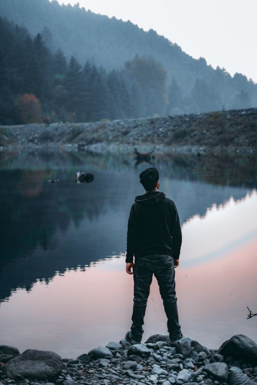 Free A lone man stands by a tranquil lake reflecting misty mountains in twilight hues. Stock Photo
