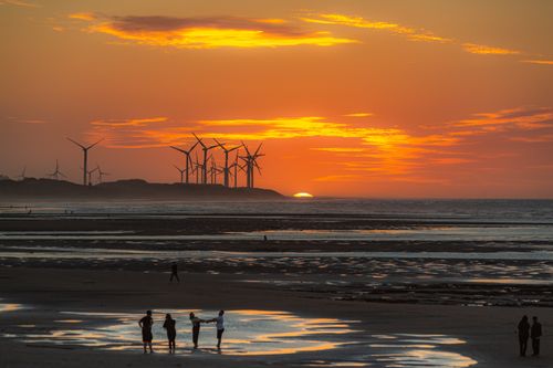 Free Silhouetted wind turbines and people on a beach at a vibrant sunset. Stock Photo