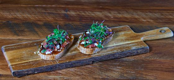 Two crostini topped with chopped tomato, red onion, crumbled feta and microgreens on a wooden paddle