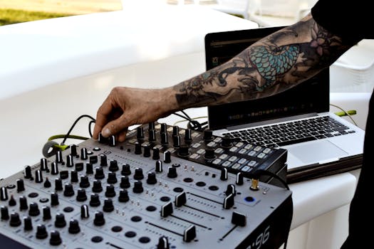 DJ with tattoos operates audio mixer and laptop outdoors. Perfect for music and lifestyle themes.