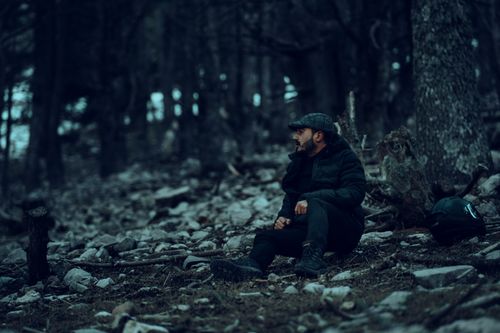 Free A bearded man in a black jacket and cap sits on the ground in a dense forest, surrounded by rocks and tree trunks. Stock Photo