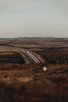 A lone car travels along a quiet road through vast brown plains under an open sky.