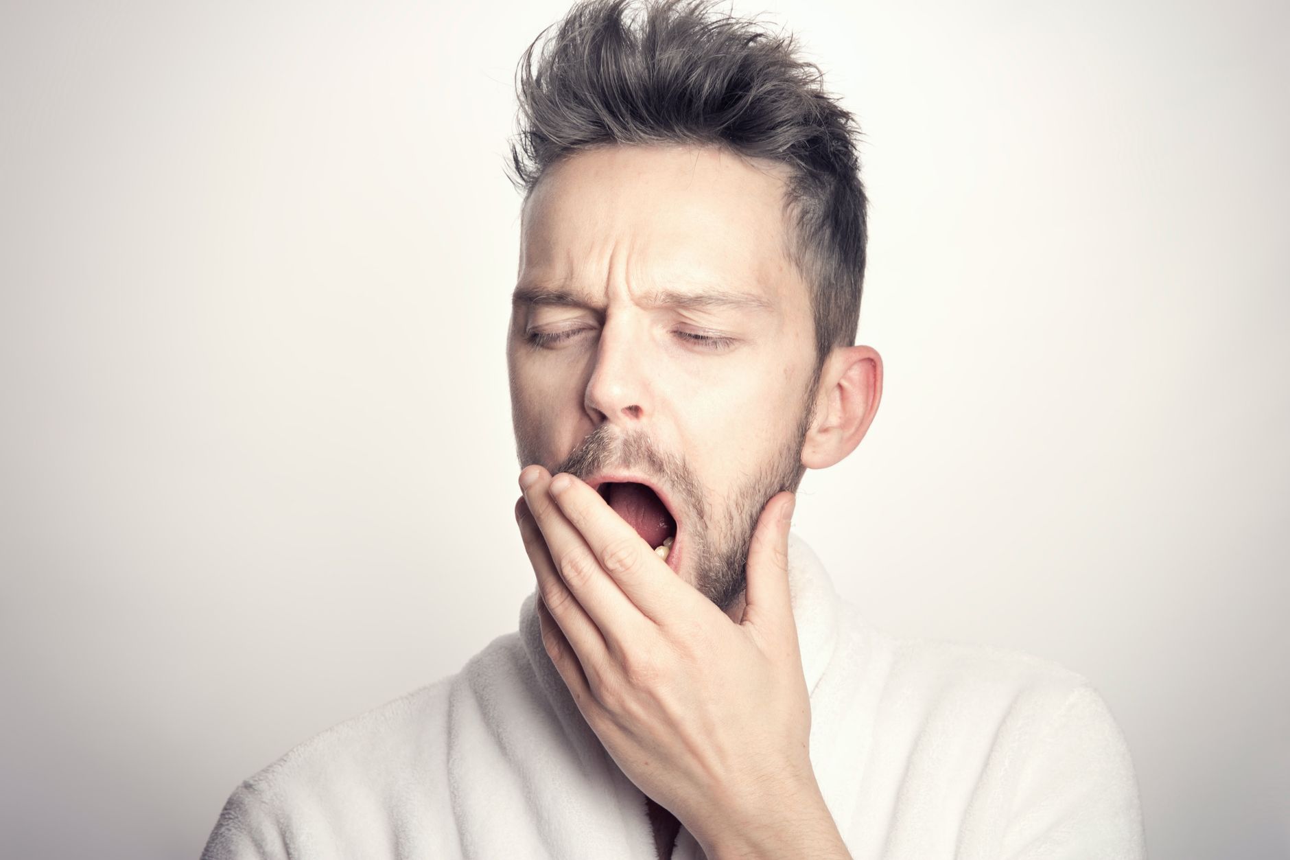 Close-up of a bearded man yawning with hand on mouth against a white background.