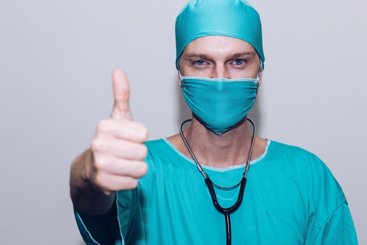A Person Wearing Blue Surgical Cap, Surgical Mask And Medical Gown Blue Doing Thumbs Up