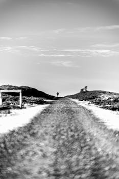 Black and white image of a solitary figure walking on a dirt road amid rolling hills.