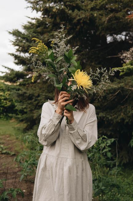 Woman in white dress holding a bouquet of wildflowers outdoors, face obscured by flowers.