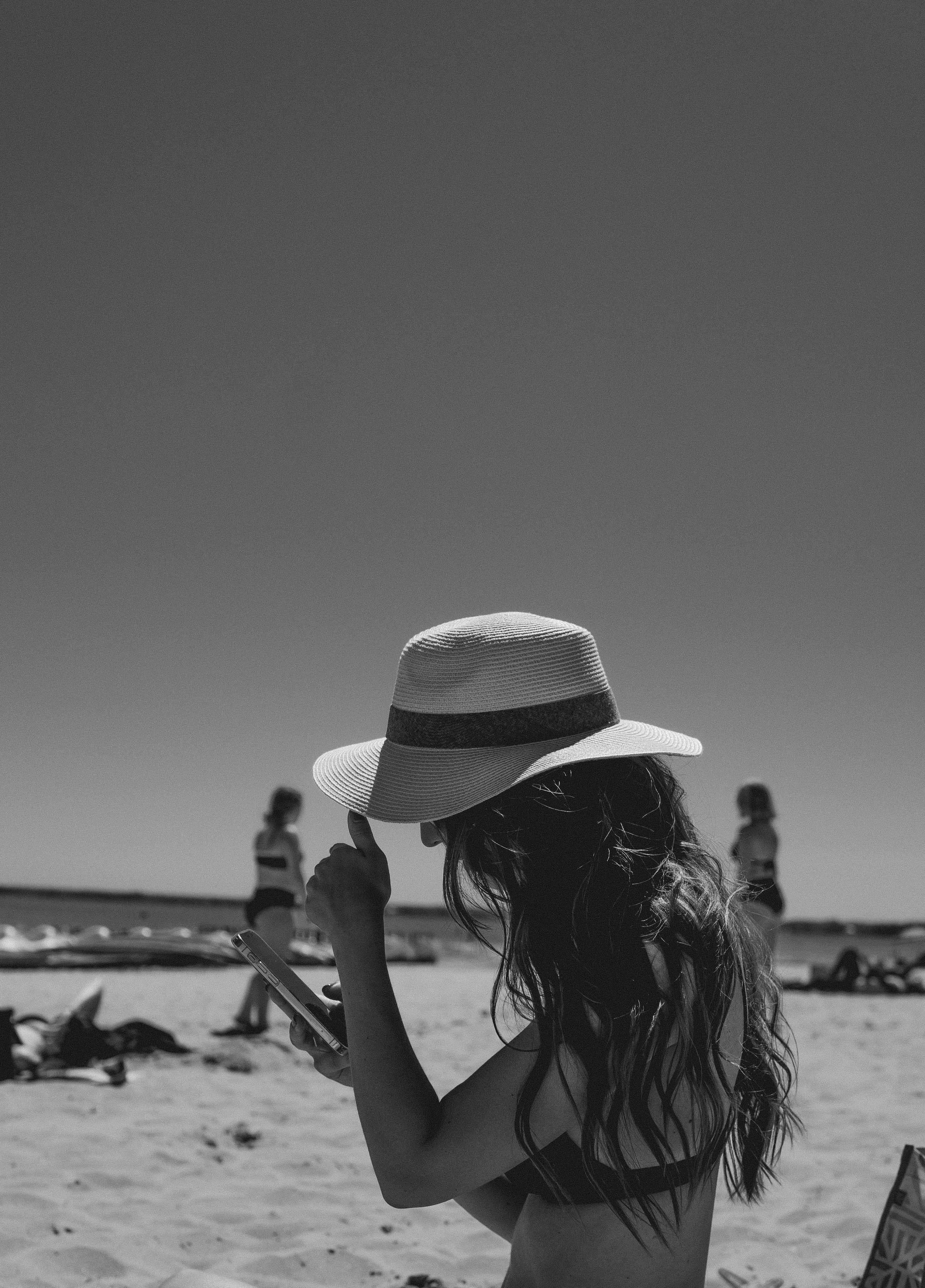 Silhouette of a woman in a fedora on a sunny beach holding a smartphone.