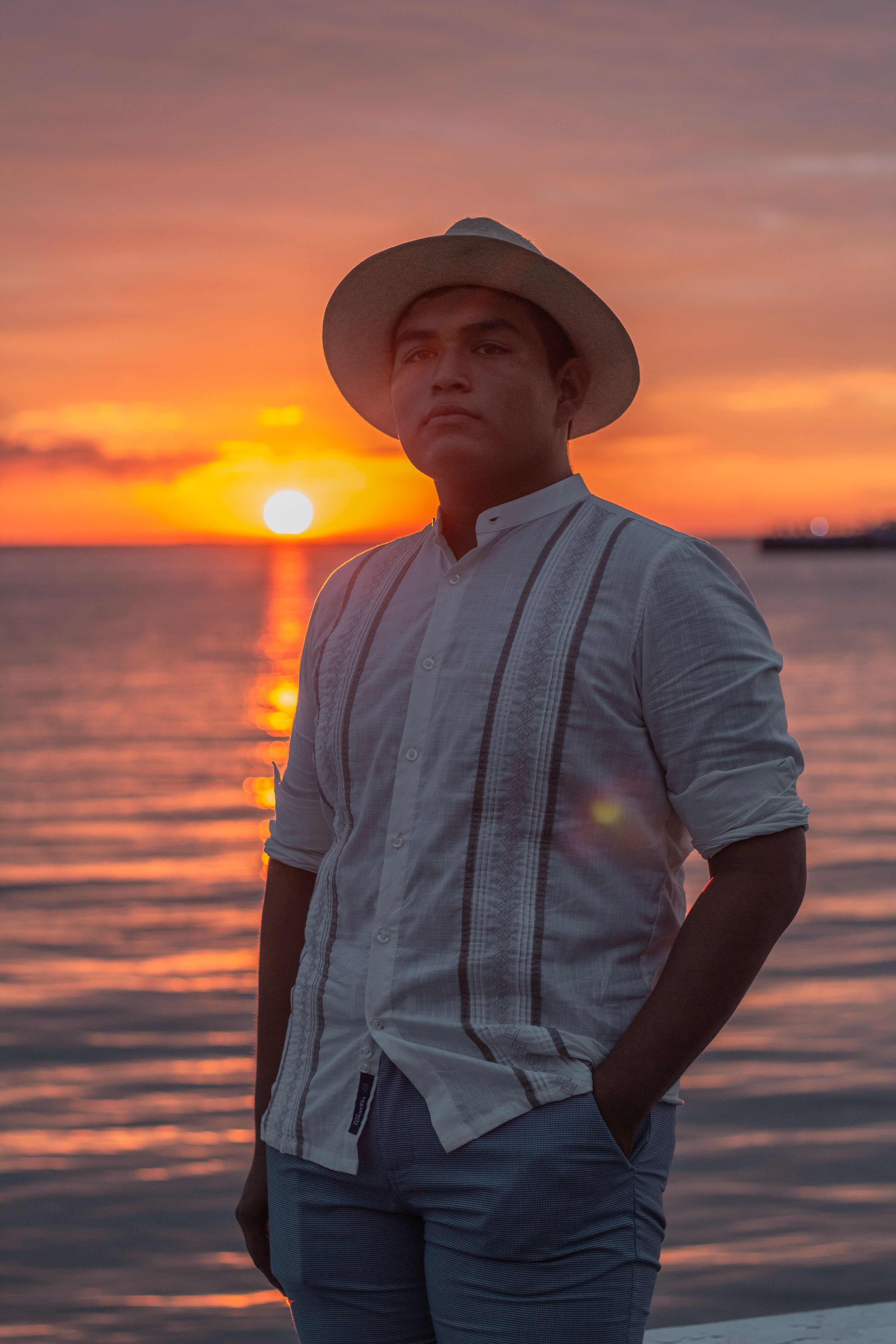 A man in a white shirt and hat stands by the ocean at sunset, hand in pocket.