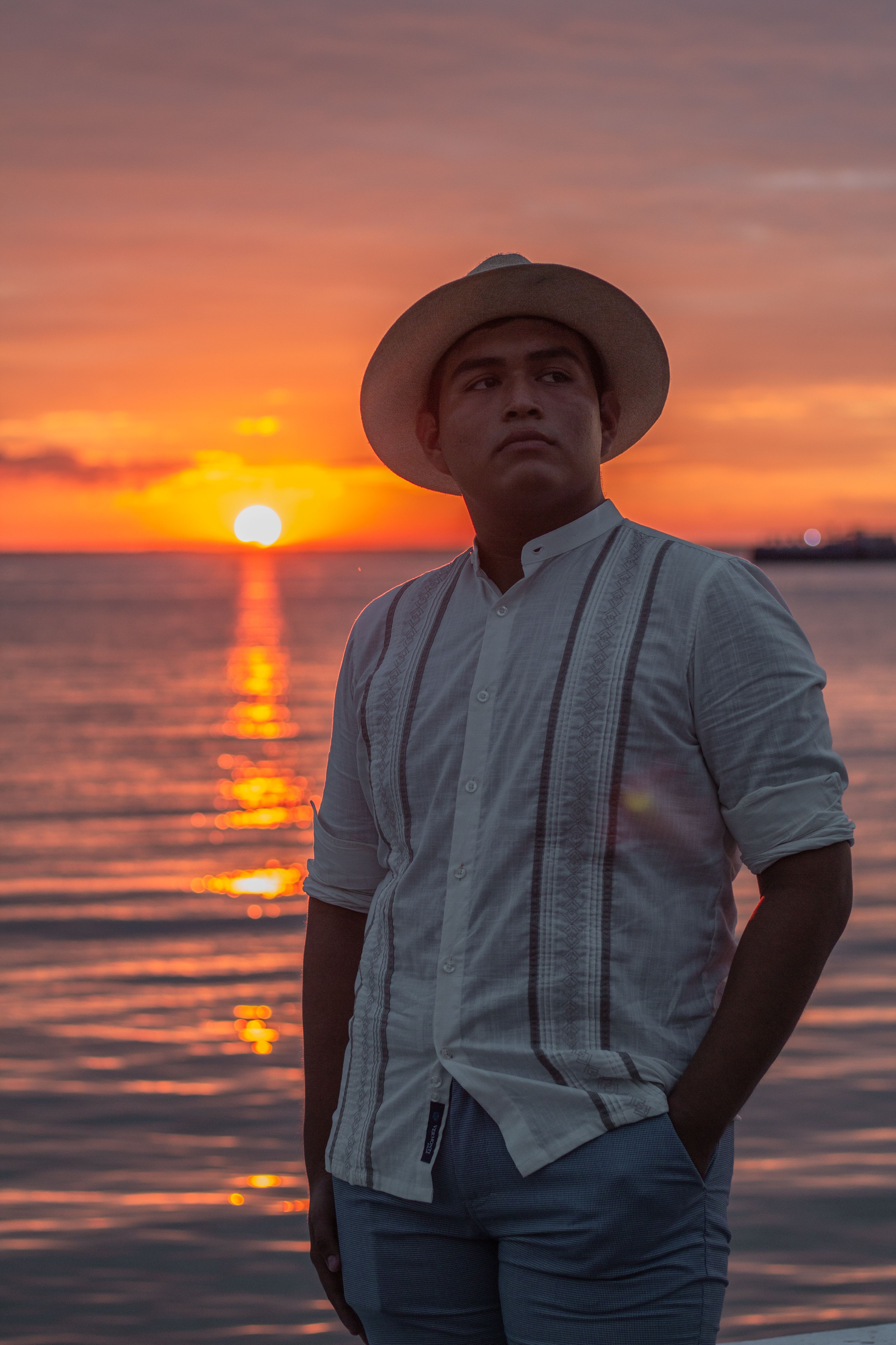 A person stands at the sea shore during a breathtaking sunset, wearing a white shirt and a stylish hat.