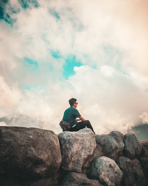 Man in Green Shirt and Black Pants Sitting on Top of Rock Cliff Under White Clouds