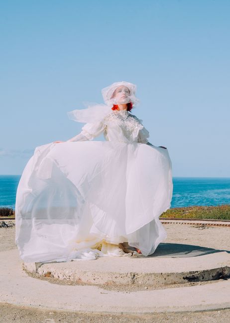 Bride in a flowing gown poses elegantly by the ocean under a clear blue sky.