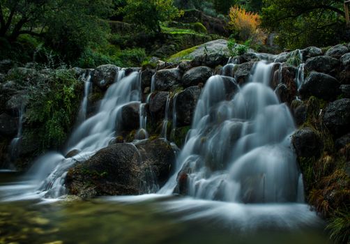 A tranquil waterfall flows over rocks surrounded by lush greenery in Covilha, Portugal.