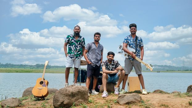 Four young musicians with instruments by a lake in Bhopal, India, enjoying a sunny day.