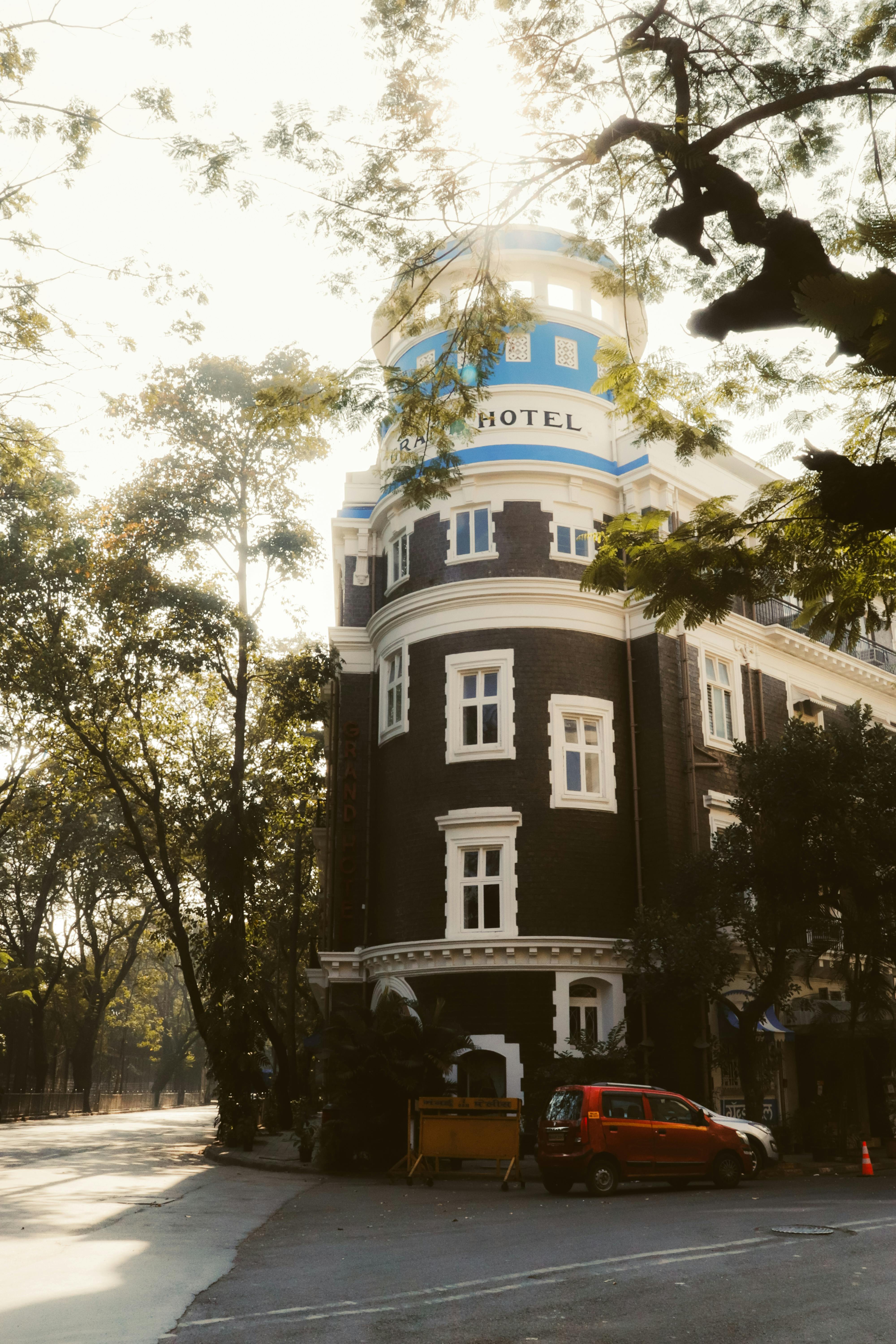 Free A historic hotel building in Mumbai surrounded by trees and parked cars on a sunny day. Stock Photo
