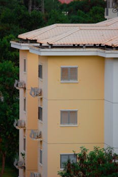 Exterior shot of a yellow apartment building with window shutters surrounded by greenery.
