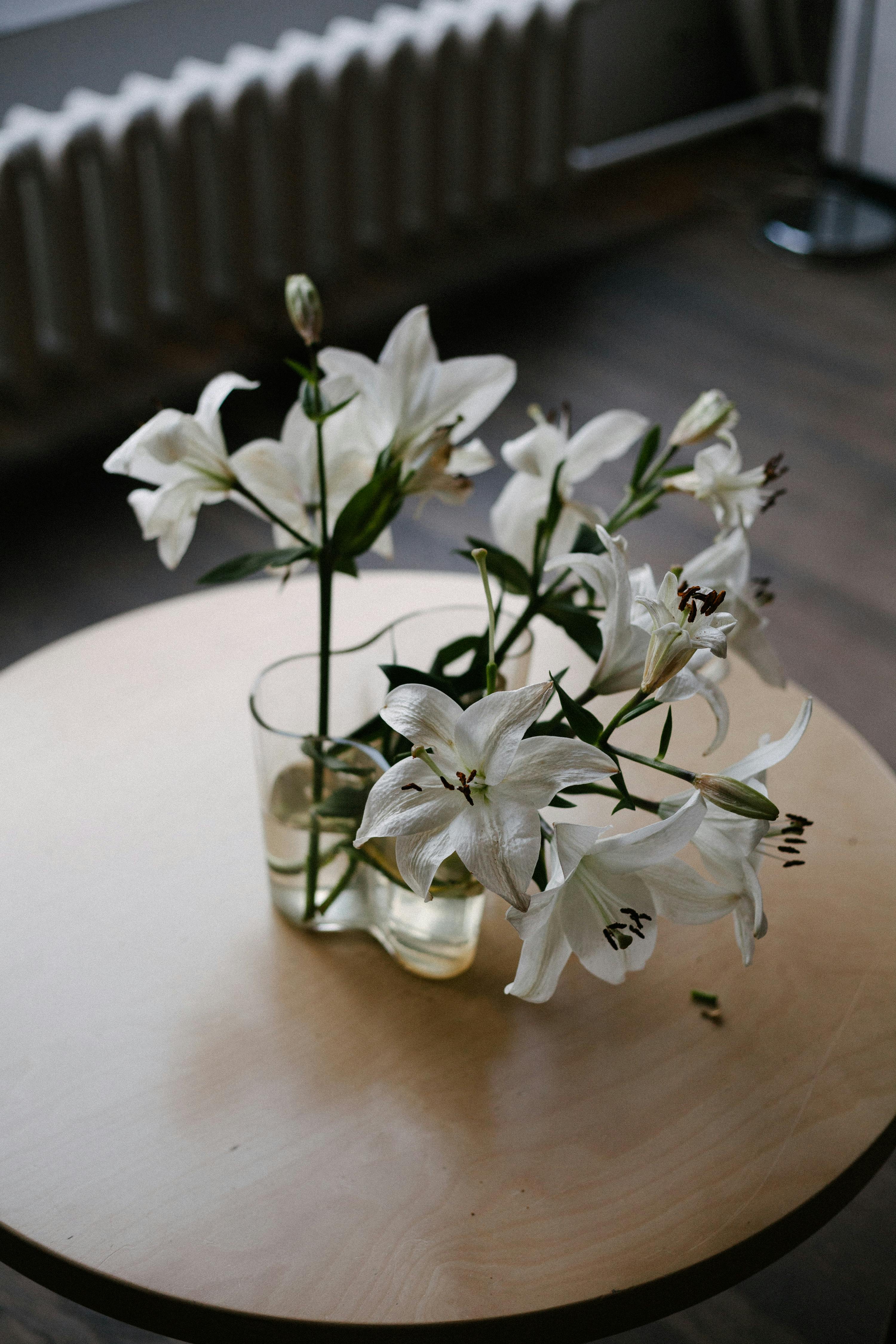 Aesthetic floral arrangement of white lilies in a glass vase on a round wooden table indoors.