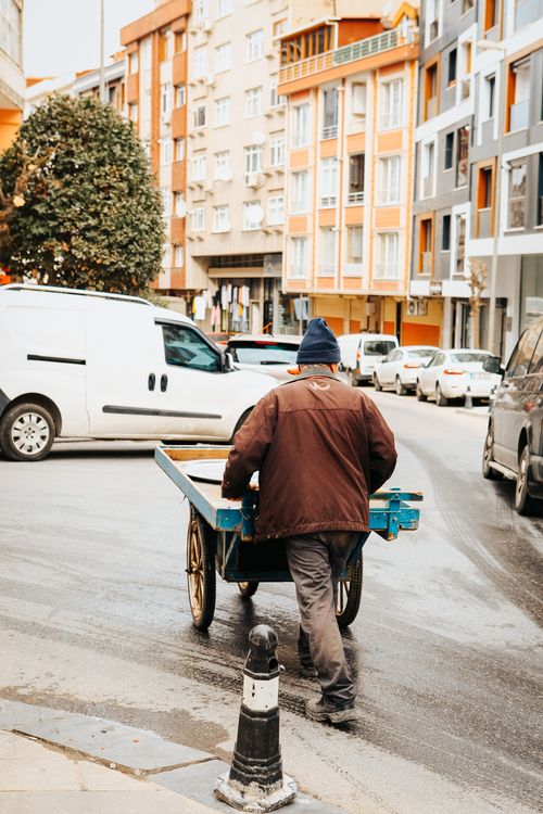 Free A man in a brown jacket pushes a cart through a busy urban street lined with colorful buildings. Stock Photo