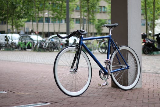 A sleek road bicycle parked against a column on a city street, surrounded by trees and buildings.