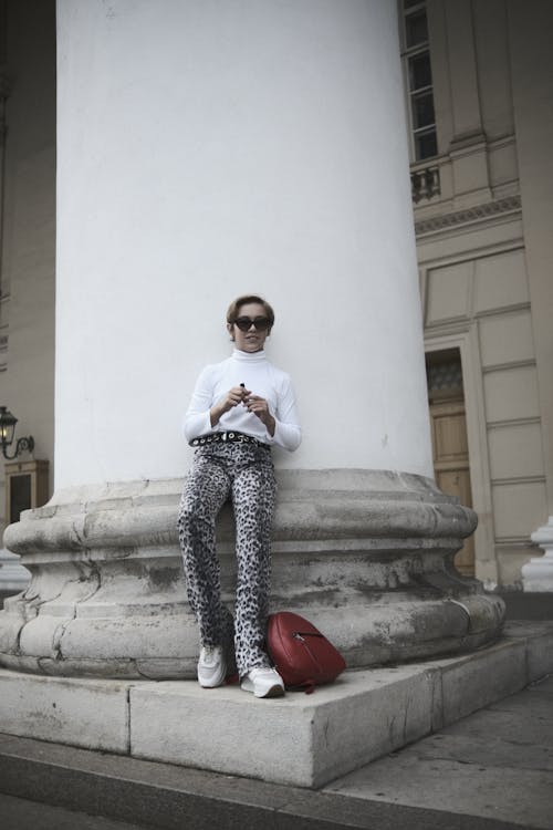 A Woman in a White Turtleneck Sweater and Animal Printed Pants Leaning on a Pillar