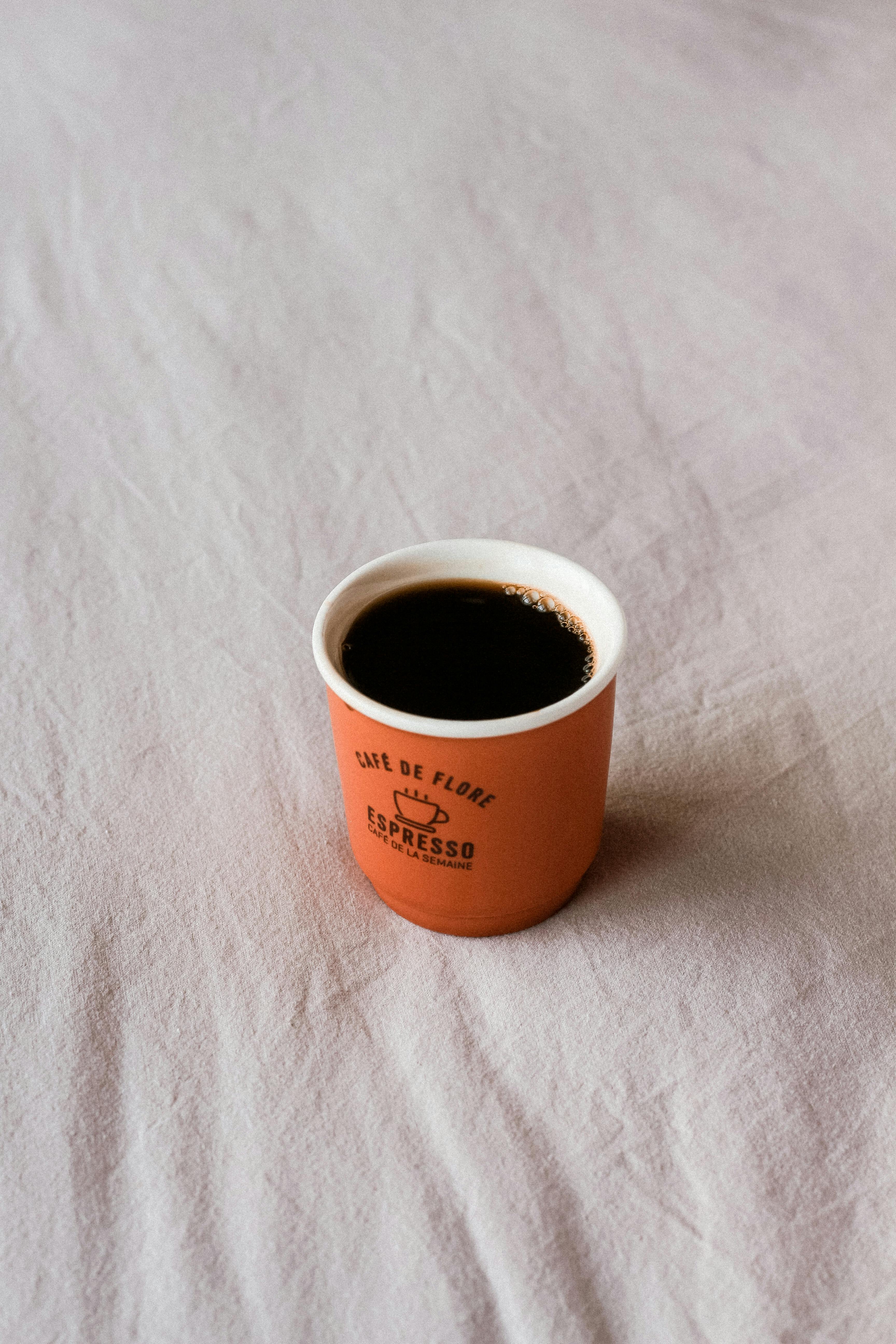 A minimalistic shot of black espresso in an orange paper cup on a light fabric surface.