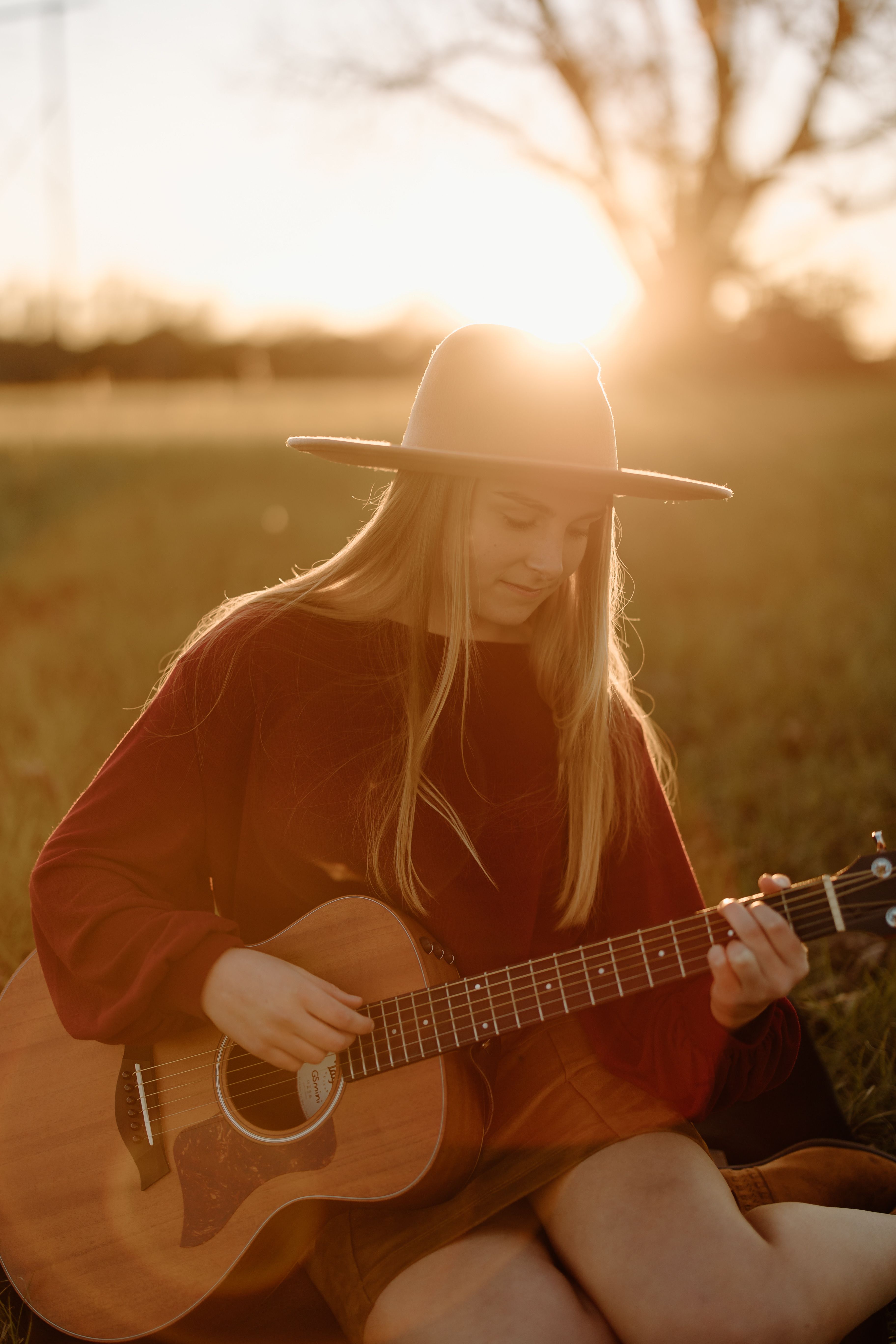 A woman in a hat plays acoustic guitar in a grassy field during a warm sunset.
