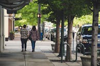 两个人走在城市的人行道上，街道两旁停着汽车，种着树木