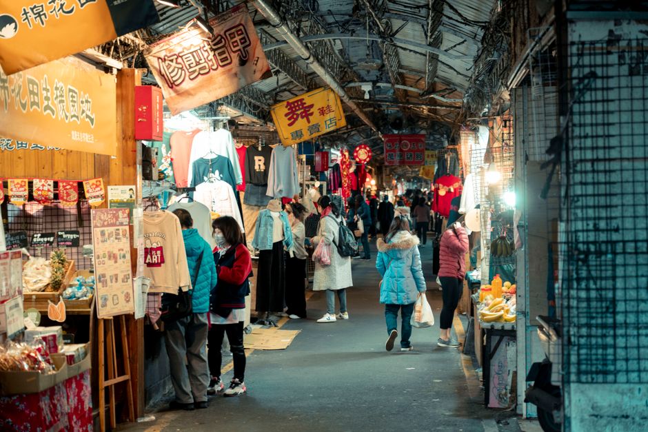 活気あるストリートマーケットのシーン、買い物客が屋台を見て回り、鮮やかなライトの下で衣服や食べ物を展示しています。