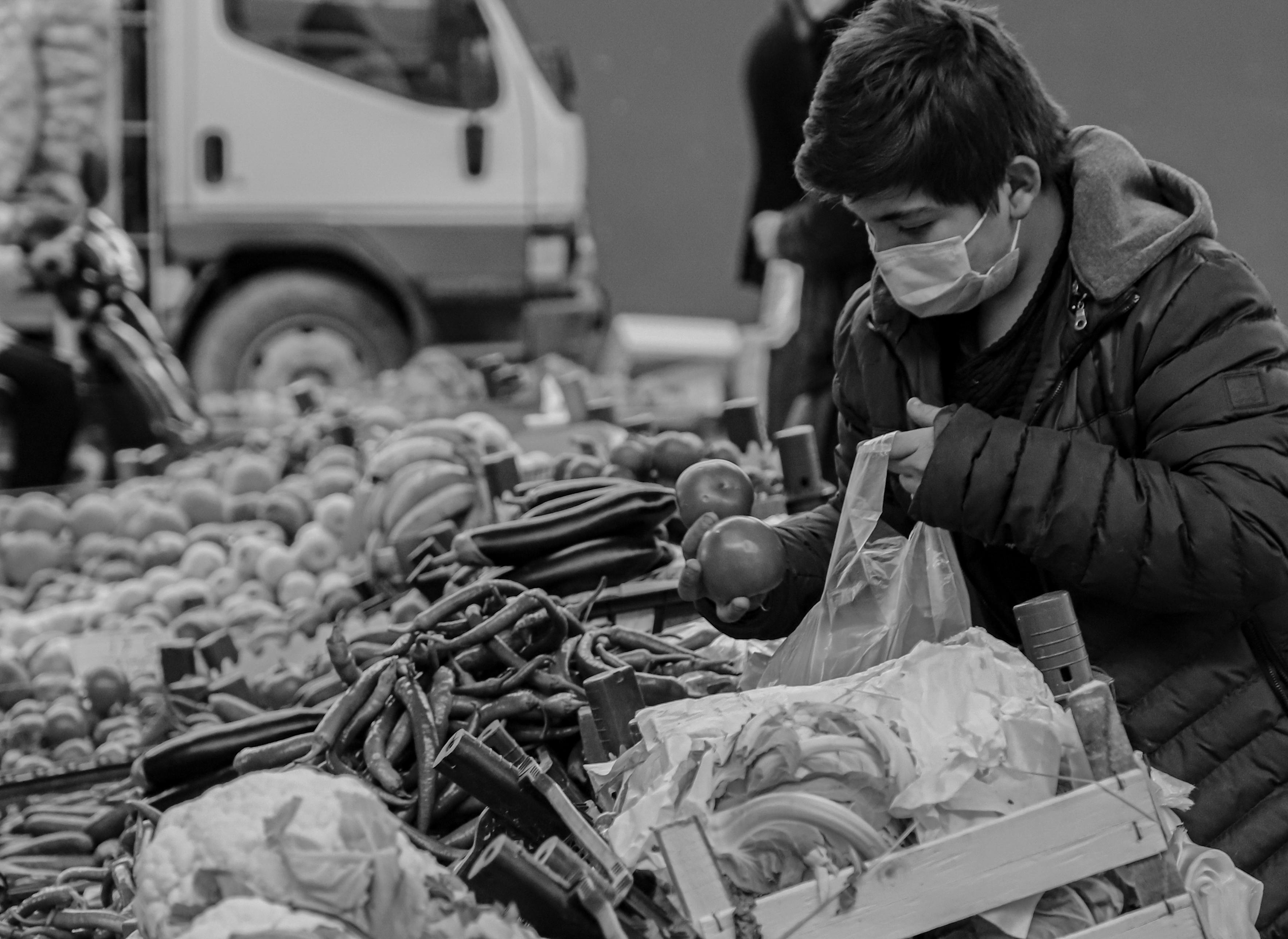 A person wearing a mask shops at a bustling vegetable market, selecting fresh produce.
