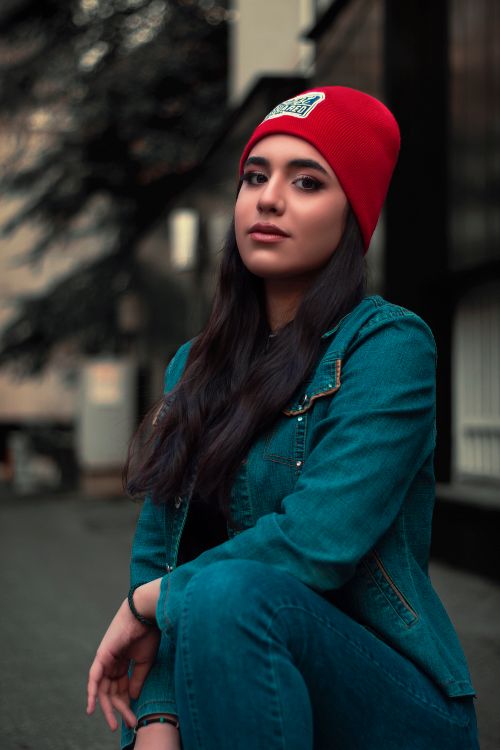 Free Confident woman in a denim jacket and red beanie posing outdoors in a street setting. Stock Photo